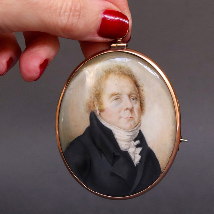 Antique Georgian era portrait miniature, hand-painted and finely detailed. Depicts a middle-aged gentleman in topcoat and cravat with cock-eyed appearance. Oval rose-gold frame with brooch and pendant fittings.