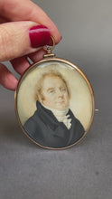 Load and play video in Gallery viewer, Antique Georgian era portrait miniature, hand-painted and finely detailed. Depicts a middle-aged gentleman in topcoat and cravat with cock-eyed appearance. Oval rose-gold frame with brooch and pendant fittings.
